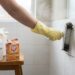 Person in yellow cleaning gloves scrubs dirty tile grout with a brush, while cleaning supplies (vinegar, baking soda, sponge) sit on a wooden stool nearby.