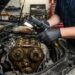 Mechanic wearing dark blue uniform and black gloves inspecting an exposed car engine with timing gears and a flashlight.