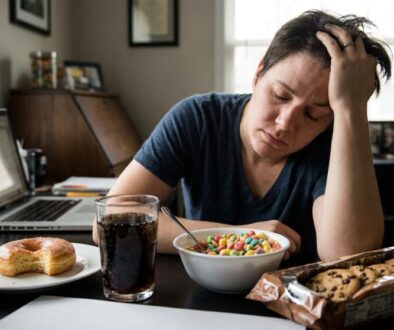 Person at a cluttered desk looks stressed, resting head in hand while eating cereal from a bowl, with a donut on a plate, a glass of soda, and a package of cookies nearby, and a laptop open in the background.