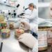 Food laboratory scene with gloved hands scooping pâté from several open canned meats on a stainless workbench; microscopes in the background.