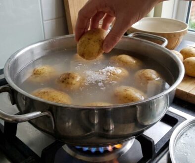 Hand dropping a potato into a pot of boiling water on a gas stove; more potatoes sit on a cutting board nearby