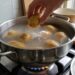Hand dropping a potato into a pot of boiling water on a gas stove; more potatoes sit on a cutting board nearby