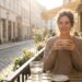 Smiling woman in a beige sweater sits at an outdoor cafe, holding a mug in the sunlight on a cobbled street.