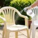 Person wearing gloves sprays a hydrogen peroxide solution on two white plastic chairs outdoors in a sunny garden.