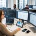 Developer seated at a desk with multiple monitors and a laptop, coding while colleagues work in an open-plan office.
