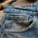 Close-up of a worn blue denim jeans pocket with copper rivets on a wooden work surface.