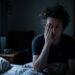 Dimly lit bedroom: a man sits up in bed with one hand covering his face, looking distressed or exhausted.