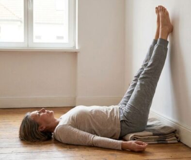 Person lying on their back on a wooden floor with legs raised and resting against a white wall, performing a legs-up-the-wall stretch on a folded blanket.