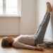 Person lying on their back on a wooden floor with legs raised and resting against a white wall, performing a legs-up-the-wall stretch on a folded blanket.