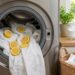 Front-loading washing machine with its door open; white towels and lemon slices inside the drum, as if for freshening or washing. On the counter are a jar and a bowl of lemons, plant, and a basket of folded towels nearby.