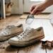 Hands sprinkle baking soda from a scoop onto a pair of dirty white sneakers on a wooden floor.