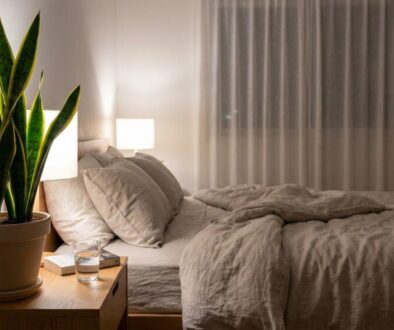 Bedroom scene with a king-sized bed, beige linens, and a tall snake plant on a wooden nightstand beside a lit table lamp.