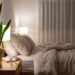 Bedroom scene with a king-sized bed, beige linens, and a tall snake plant on a wooden nightstand beside a lit table lamp.