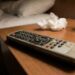 Dusty TV remote on a wooden bedside table with a glass of water and crumpled tissue, bed blurred in the background.