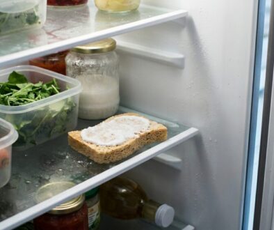 Open refrigerator shelf with containers of greens and vegetables, a jar of sauce, and a slice of bread with spread on a ice-coated shelf.