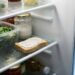 Open refrigerator shelf with containers of greens and vegetables, a jar of sauce, and a slice of bread with spread on a ice-coated shelf.