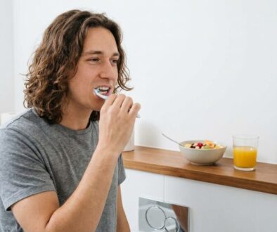 Man brushing his teeth at a wooden bathroom counter with a bowl of fruit, orange juice, and a small clock in the background.