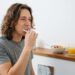 Man brushing his teeth at a wooden bathroom counter with a bowl of fruit, orange juice, and a small clock in the background.