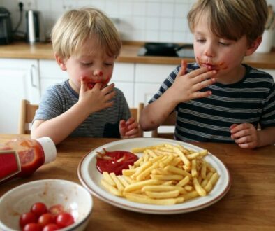 Two young boys with ketchup around their mouths eat french fries at a wooden kitchen table.