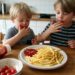 Two young boys with ketchup around their mouths eat french fries at a wooden kitchen table.