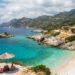 Turquoise cove with a sandy beach, thatched umbrellas, and sunbathers along a rocky coastline.