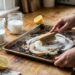 Person scrubs a greasy baking sheet with a brush, white foamy cleaner on the tray, lemons and sugar jars on a wooden counter nearby.